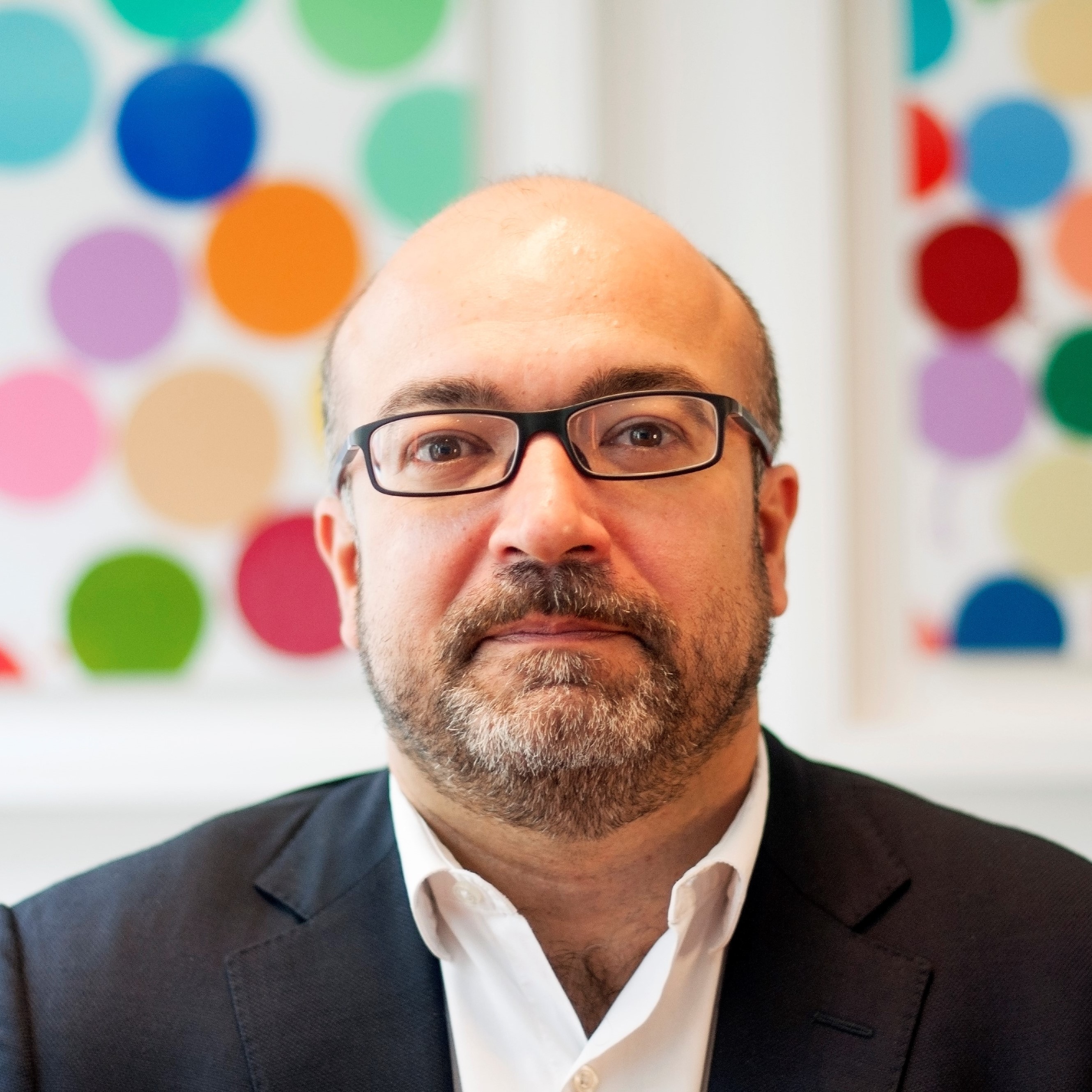 Headshot of white man with glasses in suit, sat in front of two panels with colourful baubles