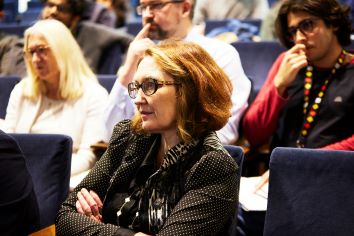 Lady in audience wearing glasses and mid-length hair, focused on speech, arms crossed