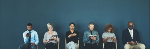 Various women and men sitting down on chairs lined up against a wall, each on their phone