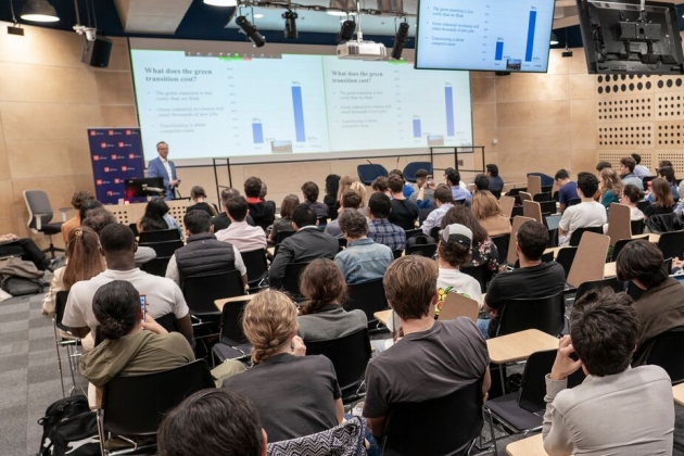 Shot from the back of a full conference room showing the speaker presenting slides