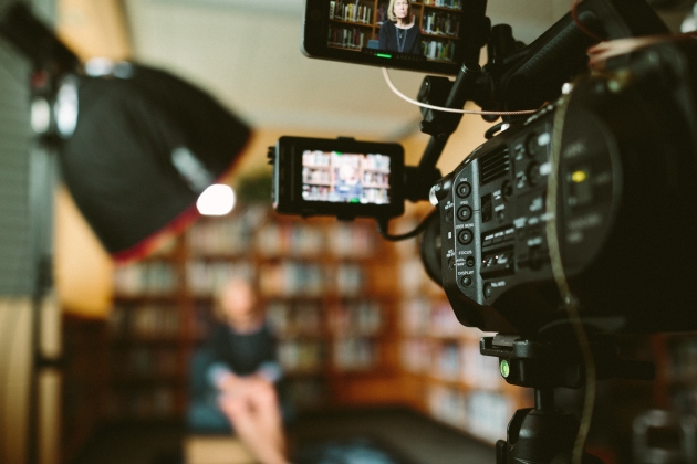 Image of a camera in the foreground, in focus, and in the background, diffuse and out of focus, a female interviewee sitting on a chair, behind a bookcase, in an office, with a light pointed at her